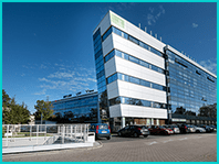 Modern multi-story office building with reflective glass facade under clear blue sky, with a parked cars and green sign on the top.